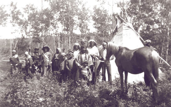 Black and white photo of 14 Cree adults and children wearing head dresses standing before a teepee in a forested setting. One man is holding a horse.