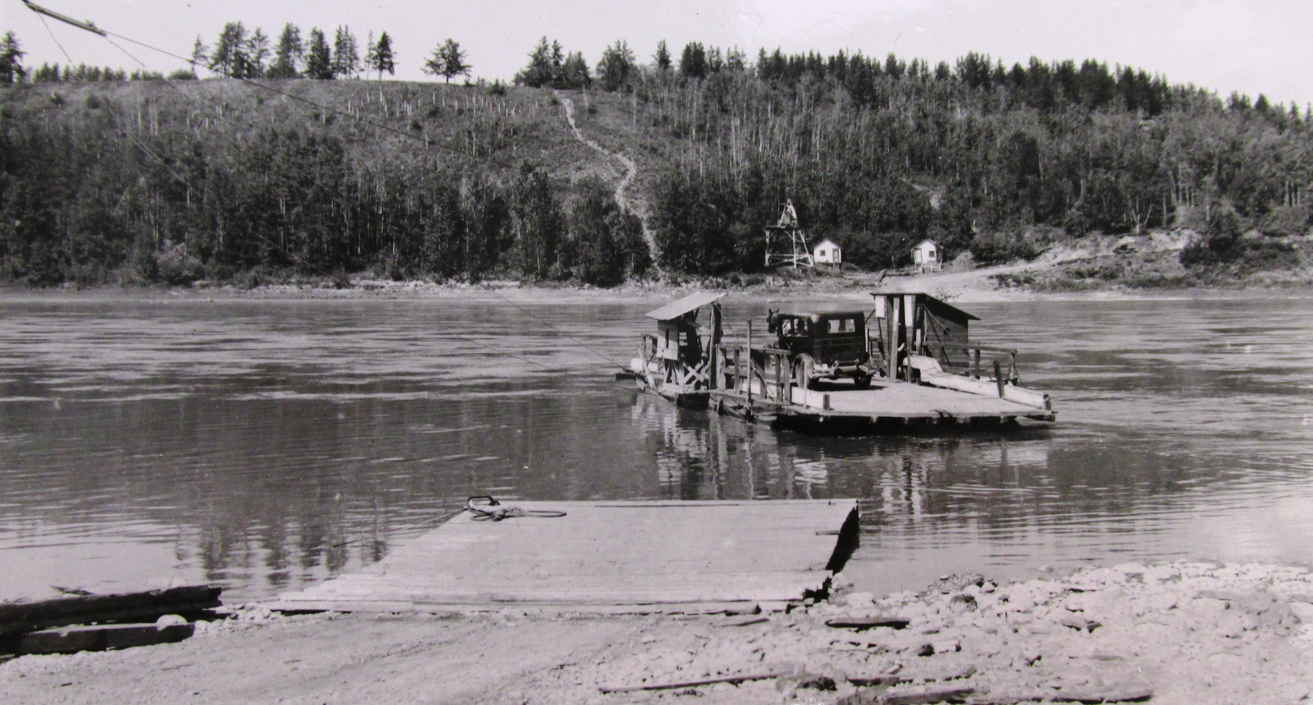A wooden ferry floating away from a dock on a river carrying a Ford Model T and with shacks built on the sides of the deck.