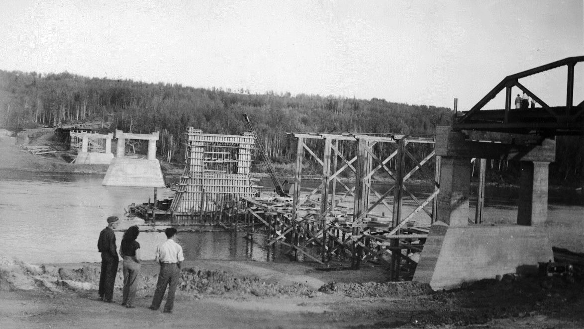 Black and white photograph taken in the early 1950s of three people standing on a river bank looking at a bridge under construction on a wide river. The are pilons in three stages on constuction; framing, metal rebar framing and poured concrete. There is no deck on the bridge.