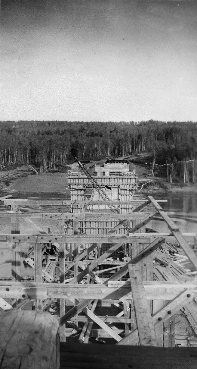 Black and white photograph of a bridge under construction across a river.