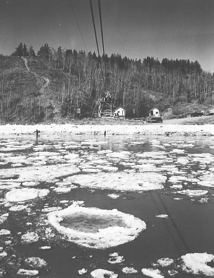 Black and white photograph taken from a cable care suspended over a river that has large chunks of ice floating in it