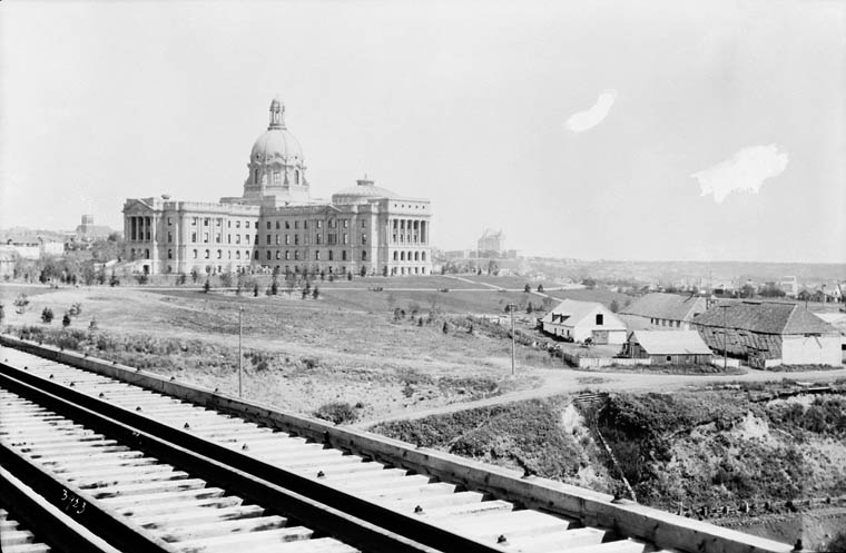 Black and white photograph of an old-fashioned, four-story government building constructed with a central dome as an architectural feature. The photo was taken from the deck of a bridge with railway tracks on it. 