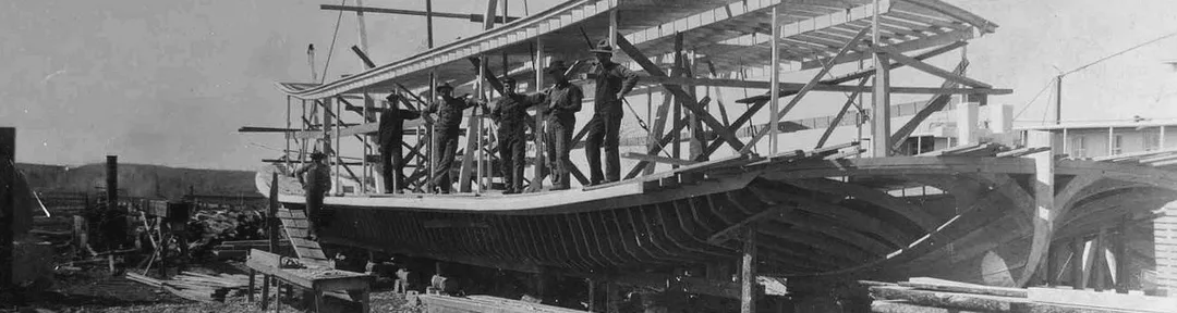 A photograph taken in 1916 showing a steamboat under construction with wooden framing only. On the deck of the boat, six men a standing, looking at the camera.