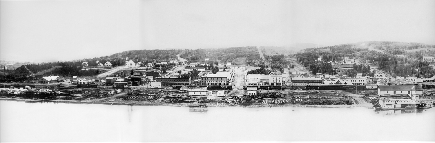 Black and white photo of a small town built up from the shore of a large river. A steamboat is landed on the riverbank where there is a laumber yard and warehouses. A train and train station can be seen, as well as two large hotels and and other smaller buildings.
